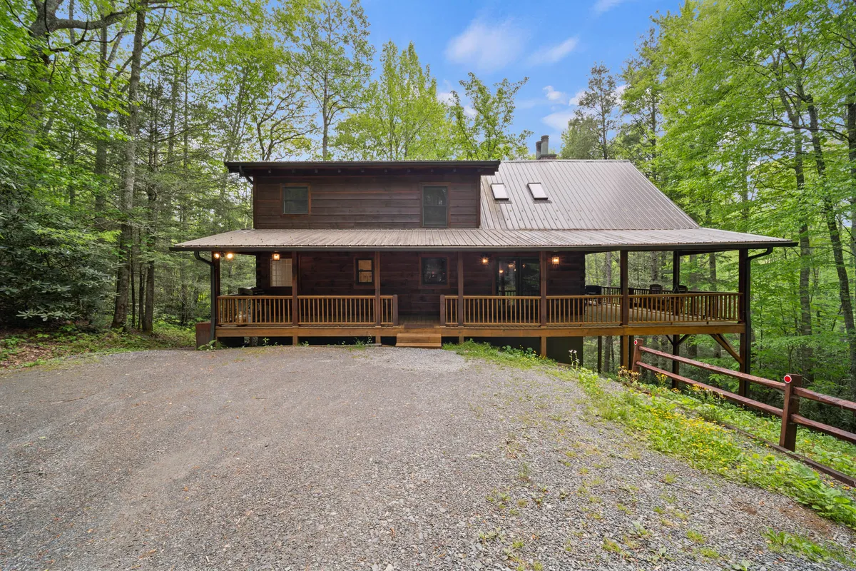 The exterior of Log Heaven, with its inviting wooden porch, large windows, and a gravel driveway set among tall trees. The porch is perfect for relaxing and offers sunset views, hinting at intimate evenings and shared outdoor experiences.