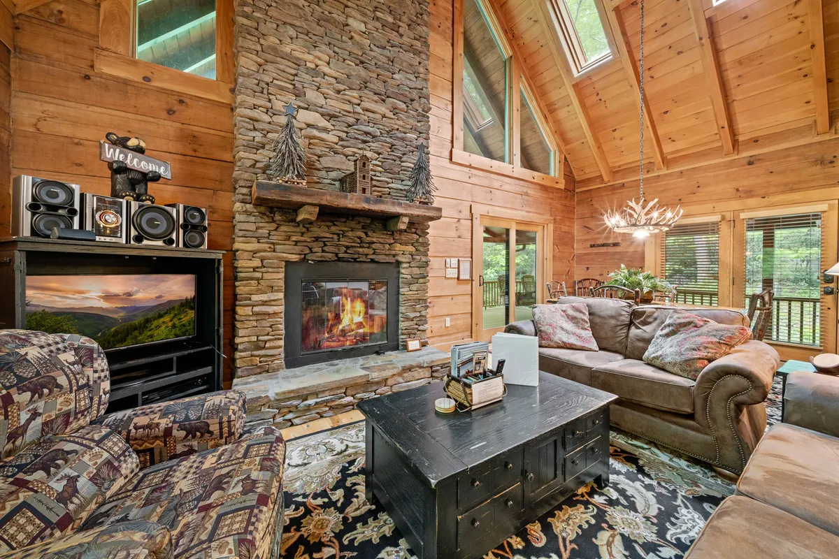 A spacious living room featuring a prominent stone fireplace set against wooden beams, with large windows allowing natural light to stream in. Cozy seating arrangements invite relaxation, and decorative lighting adds a warm, welcoming ambiance. The setting is clearly designed for gatherings, comfort, and fireside enjoyment.