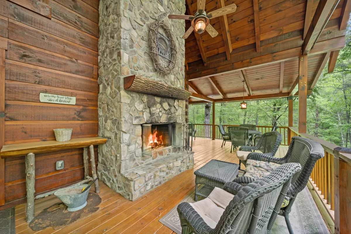 A cozy outdoor stone fireplace under a wooden ceiling with comfortable woven chairs arranged around it. Decorative accents like a seasonal wreath add warmth and charm, while the view beyond hints at lush nature. The scene feels welcoming and ideal for social gatherings or quiet fireside moments, year-round.
