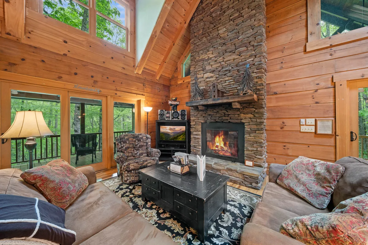 A well-appointed living area with a stone fireplace, large windows overlooking lush greenery, comfortable couches, and a dark coffee table. The arrangement creates a setting ideal for conversations, relaxation, and enjoying nature from indoors.
