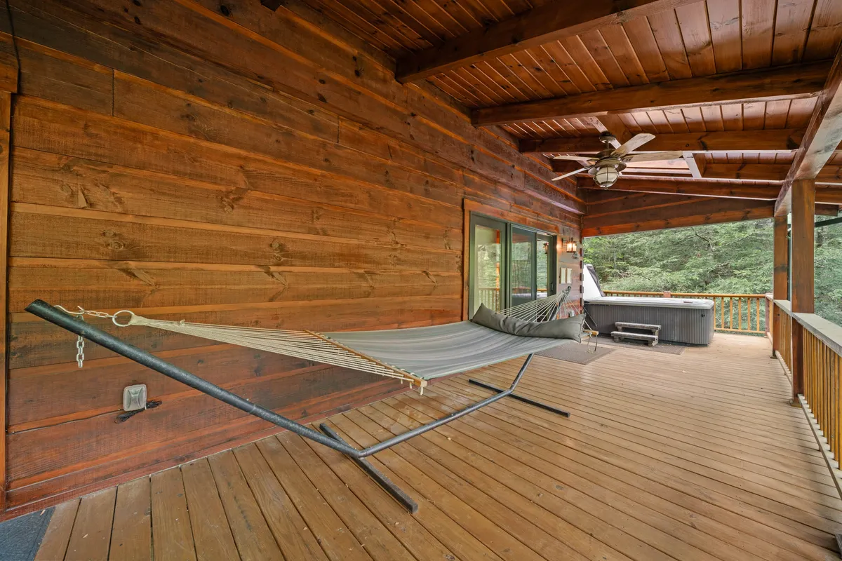 This exterior image features a serene wooden deck equipped with a hot tub, a hammock, and comfortable seating, all overlooking lush greenery. The setting is peaceful and private, with a ceiling fan hinting at year-round comfort. The inviting deck offers opportunities for stargazing, soaking in the hot tub, and cozy moments outdoors.