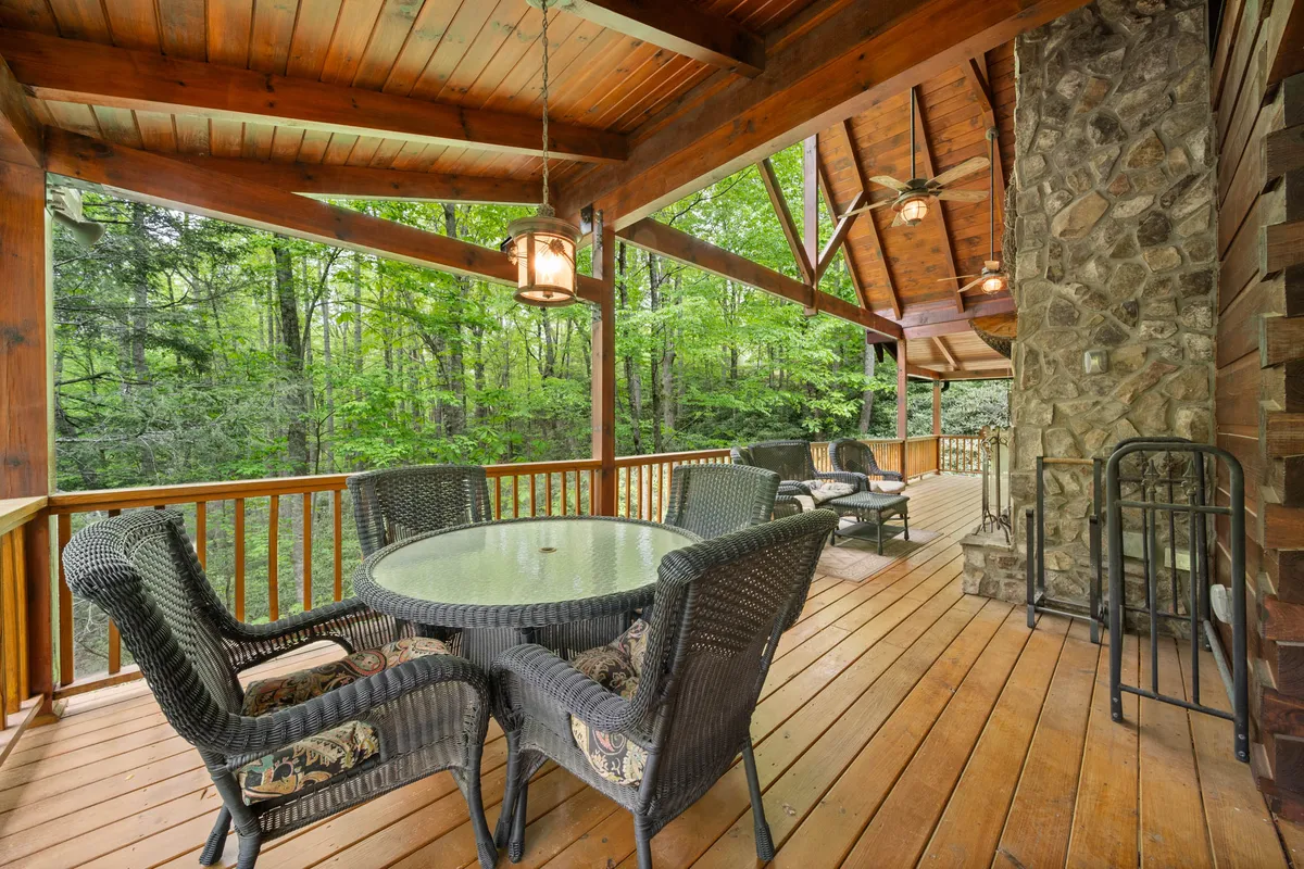 A spacious wooden deck furnished with a glass table, comfortable seating, and surrounded by lush greenery. The stone chimney hints at a cozy fireplace inside, while the deck provides a seamless transition between luxury indoor comfort and the beauty of the outdoors.