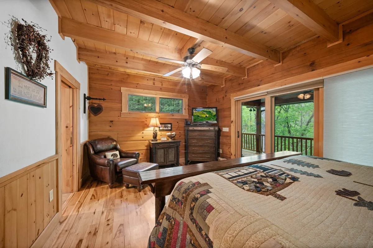 A warmly lit bedroom with wooden beams overhead, a cozy quilted bedspread, an armchair beside a lamp, and a television set. Large windows allow natural light and offer a glimpse of nature outside, creating a space that feels both restful and inviting.