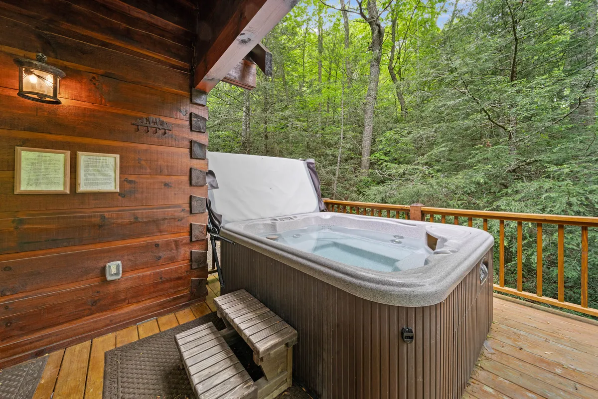 A bubbling hot tub sits on a spacious wooden deck surrounded by tall trees. The deck overlooks lush greenery, and the tranquil atmosphere is enhanced by the natural beauty of the surrounding forest. Soft evening light suggests the opportunity for sunset watching or stargazing from the comfort of the hot tub.