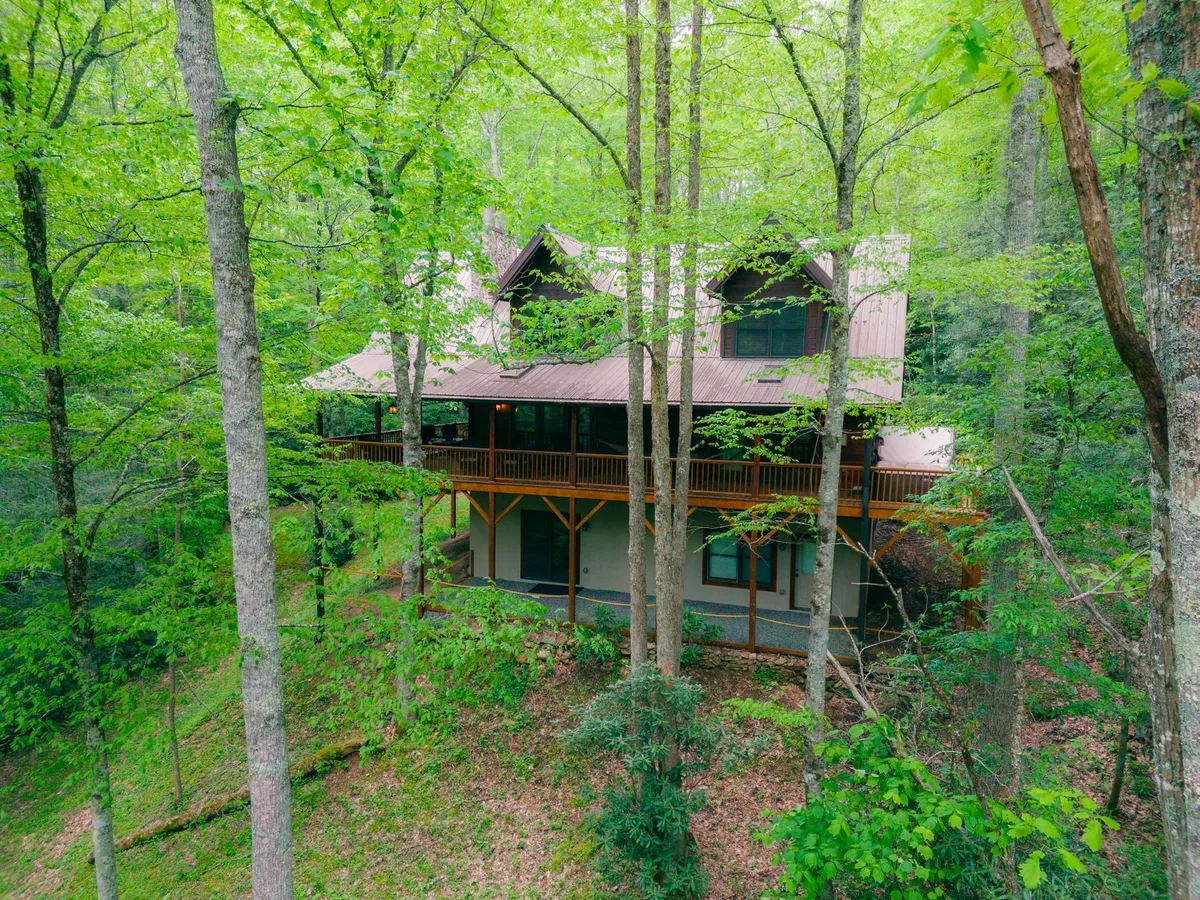 A wide shot of the cabin nestled among tall green trees, showcasing a spacious deck that overlooks the forest. The setting feels both secluded and inviting, suggesting opportunities for relaxing on the deck, exploring nearby trails, or gathering for evening fun in a peaceful mountain environment.