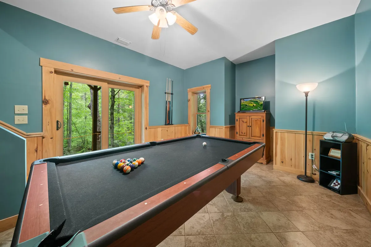 A cozy, sunlight-filled game room features a pool table, large windows with nature views, wooden cabinetry, and a warm floor lamp. The space radiates energy and fun, suggesting family tournaments, casual relaxation, and lively game nights indoors after a day outside.