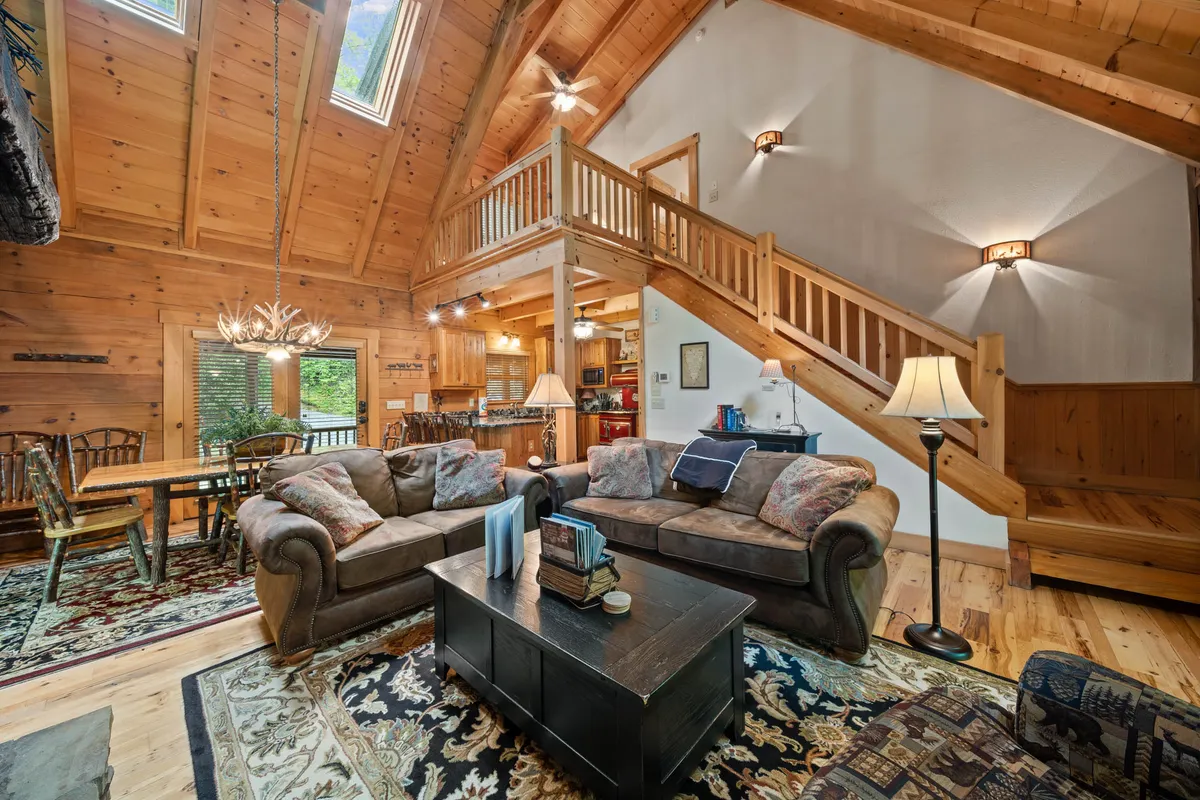 The image showcases the inviting interior of a luxury log cabin, with a plush sofa set, rustic coffee table, cozy dining area, a striking wooden staircase, and a warm chandelier casting a golden glow. The space radiates comfort and charm, encouraging gatherings, relaxation, and shared moments.