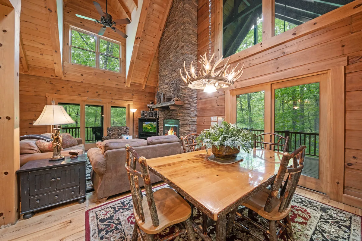 A rustic dining area with a large wooden table, antler chandelier, and expansive windows overlooking the forest. The stone fireplace adds a cozy ambiance perfect for gathering, sharing meals, and making memories.