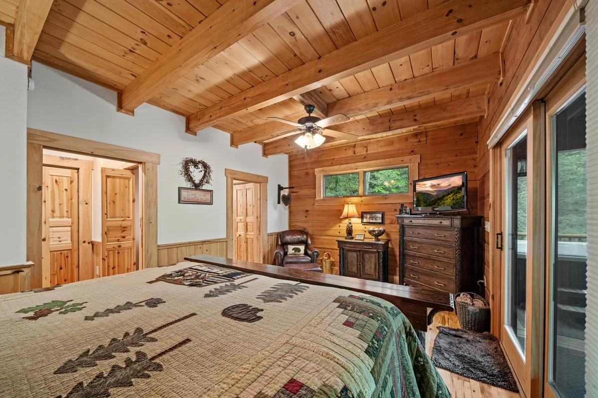 A welcoming bedroom with a soft quilted bedspread, polished wooden furniture, a lamp for reading, and a window offering picturesque forest views. The serene atmosphere is perfect for a restful night's sleep or a quiet afternoon with a book.