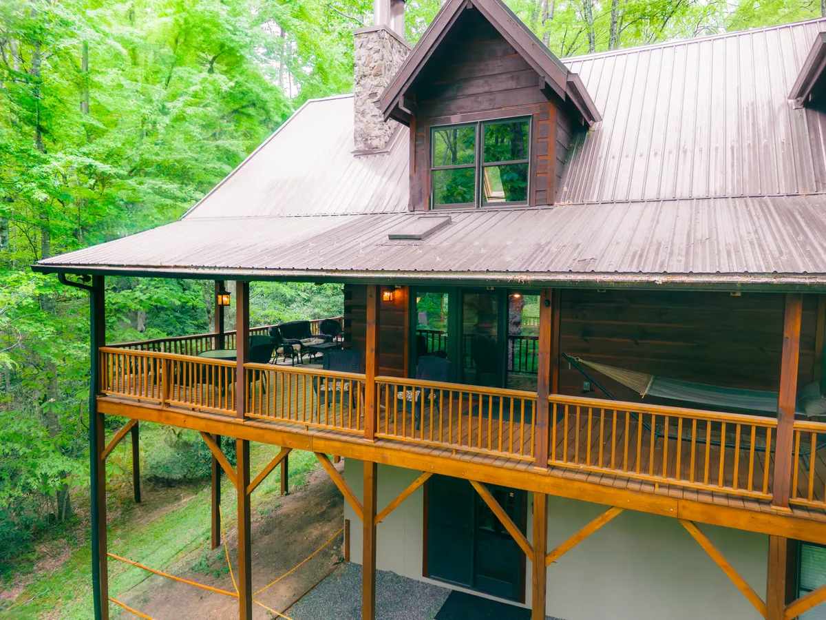A broad wooden deck wraps around the cabin, featuring a prominent stone chimney, large windows reflecting surrounding greenery, and cozy seating. The deck is bathed in natural light, ideal for lounging or sharing meals outdoors while embracing the forested setting.